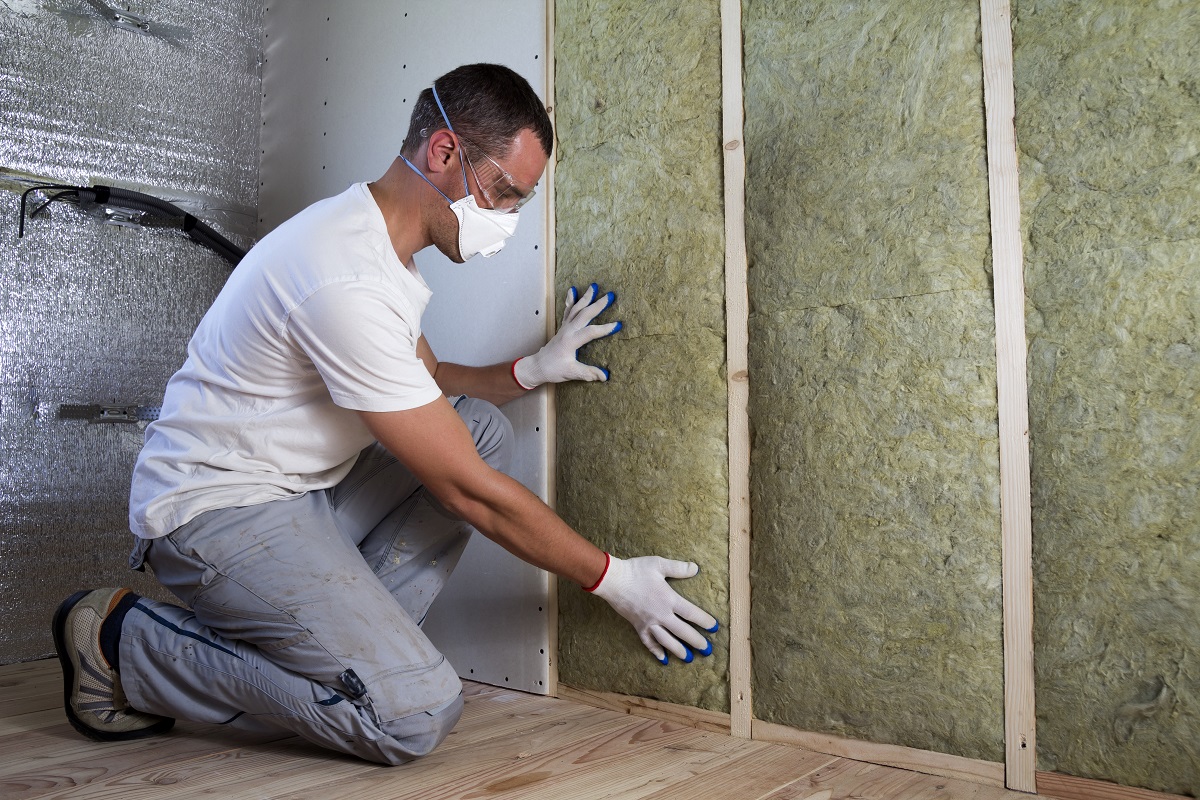 Человек в белой футболке и серых штанах устанавливает каменную вату РОКВУЛ на стену.